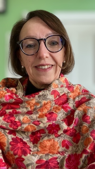A person with short, straight brown hair is smiling at the camera. They are wearing large round glasses, pearl earrings, and a colorful shawl with red, orange, and pink floral embroidery. The background features a green wall and a white door.