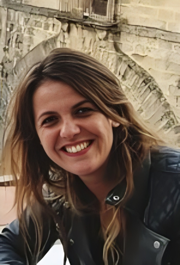 A smiling person with shoulder-length brown hair wearing a dark jacket outdoors in front of a stone archway.