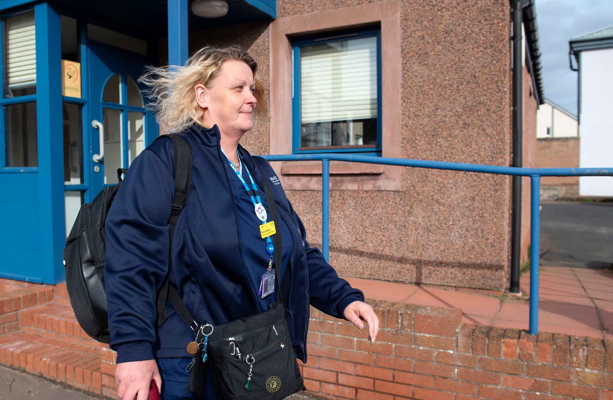 A person with wavy light-coloured hair walking outside a building in the sun. They wear a dark blue jacket with a lanyard visible that has several badges attached. They carry two black bags, a rucksack on one shoulder and a small bag across the front of their body, and a red flask. In the background a brick building with blue-framed window and doors and a brick disability ramp.