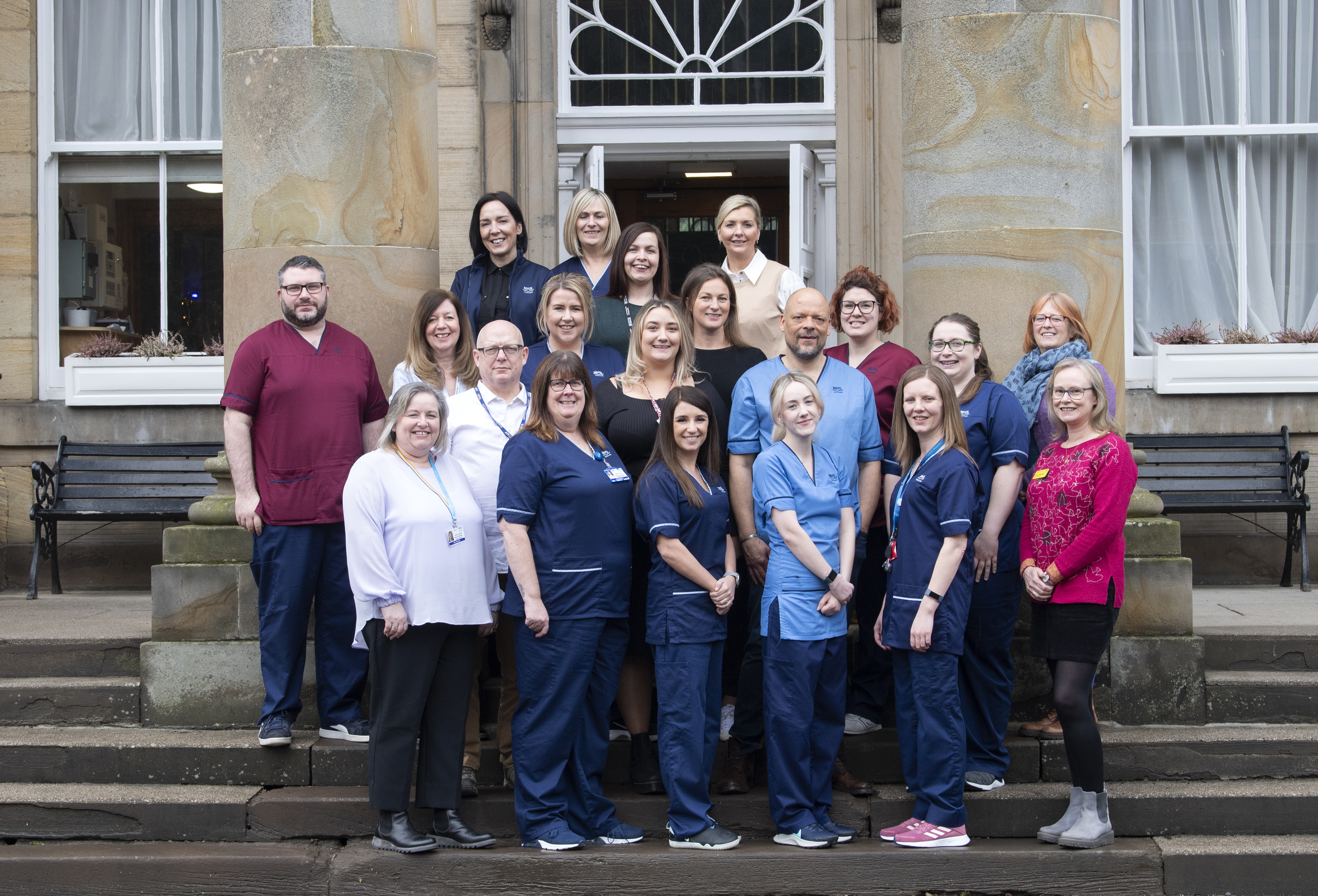 A group of 20 people pose together on the steps of a stone building with large columns and tall windows. The group includes individuals dressed in medical uniforms of various colours — mostly navy, light blue, and burgundy — as well as others in casual or business attire. They are arranged in multiple rows and are smiling at the camera. The setting appears to be outside a historic or institutional building, and benches are visible on either side of the group.