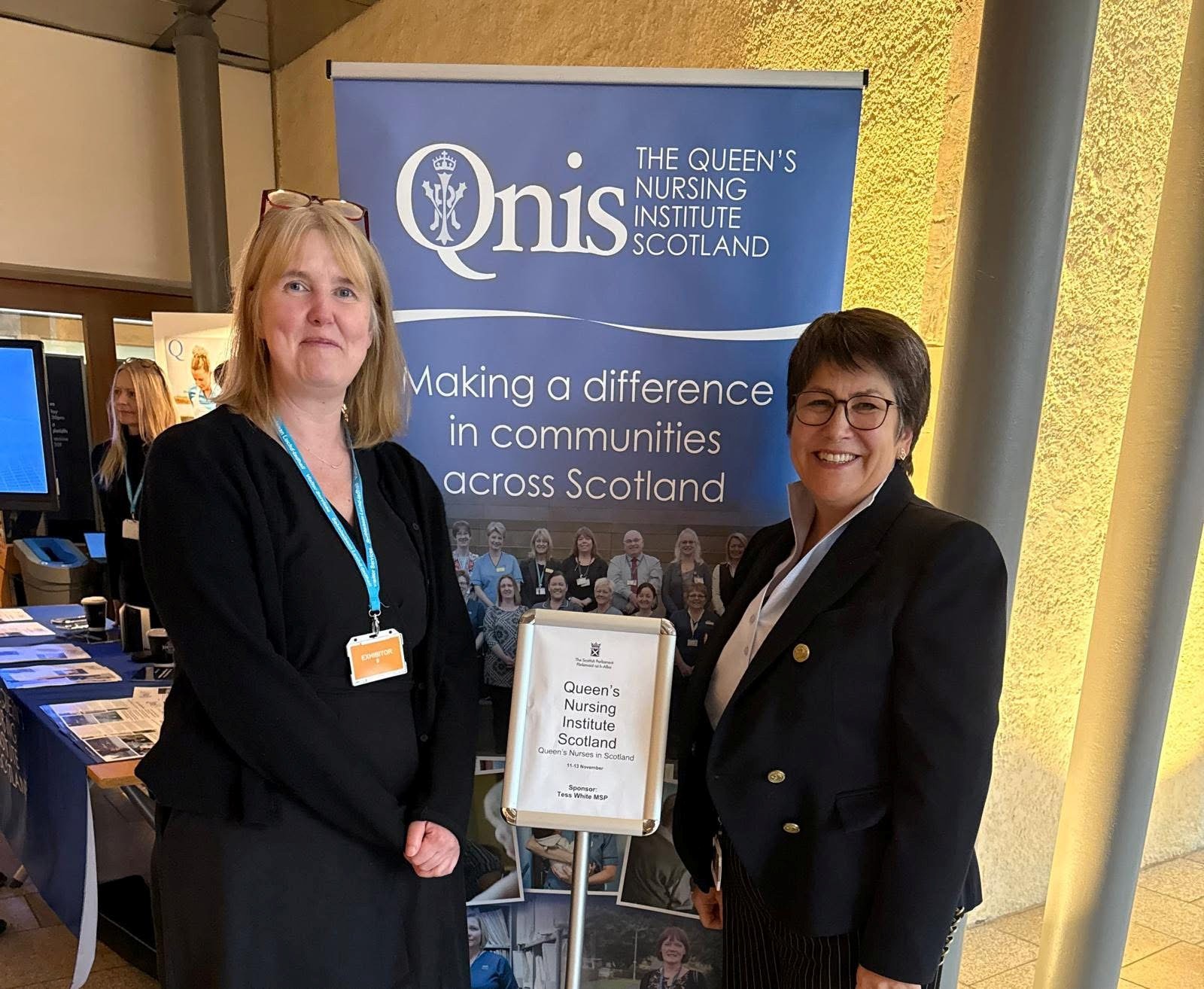 Two people stand smiling in front of a display for the Queen’s Nursing Institute Scotland. One person on the left wears a dark outfit with a light-blue lanyard, and the person on the right wears a dark blazer and glasses. Behind them is a blue banner with the organisation’s name and the text “Making a difference in communities across Scotland.” A stand with an information sign and additional display tables are visible in the background.