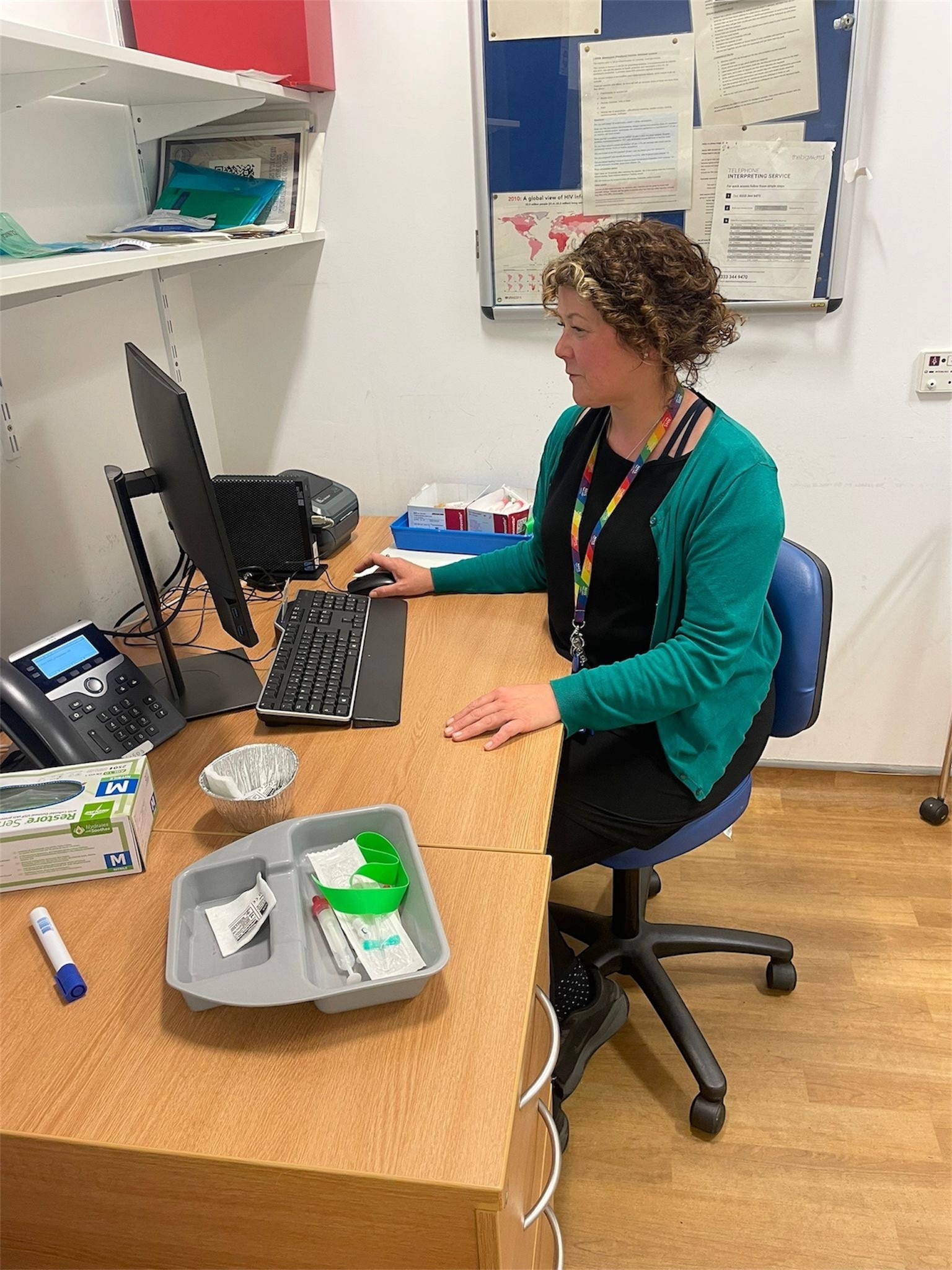 A person with short curly hair sits at a desk in an office, working on a desktop computer. They are wearing a green cardigan over dark clothing and a rainbow lanyard. On the desk are a phone, a keyboard, a medical tray with supplies, a box of gloves, a marker, and other office items. Behind them, a bulletin board with papers is mounted on the wall.