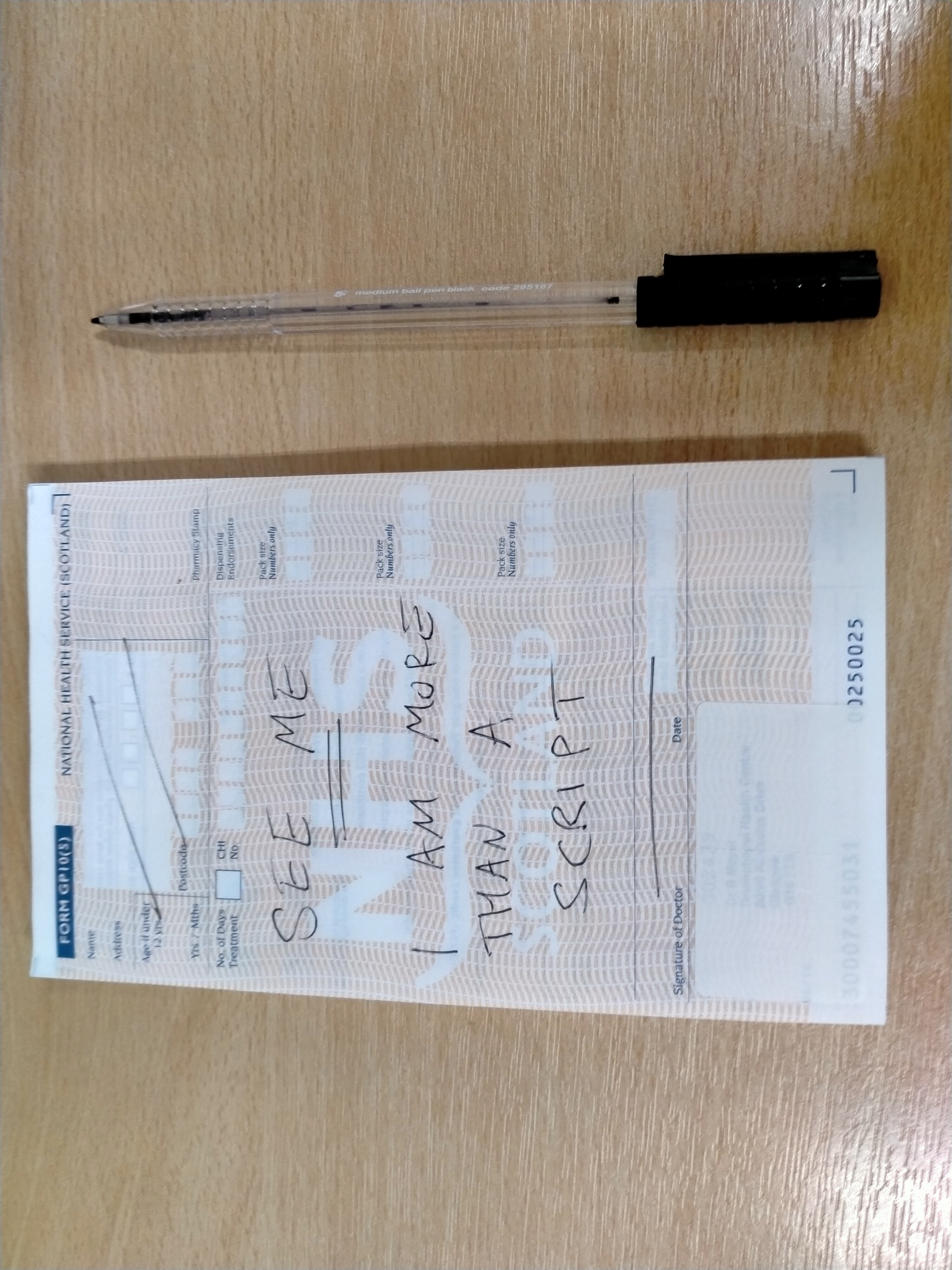 The image shows a prescription with NHS Scotland in white text across the centre and with the words See me I am more than a script written across it in black ink. A black pen sits to the right of the sheet.
