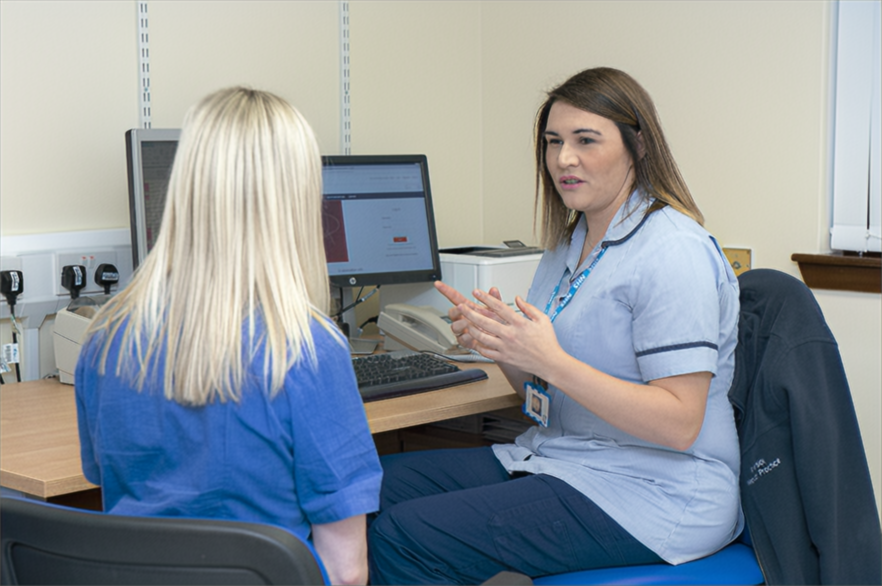 A healthcare worker in a clinical setting speaks with another person seated across from them. Both are wearing blue uniforms, one has long brown hair, the other has long blonde hair, and a computer is visible on the desk between them.