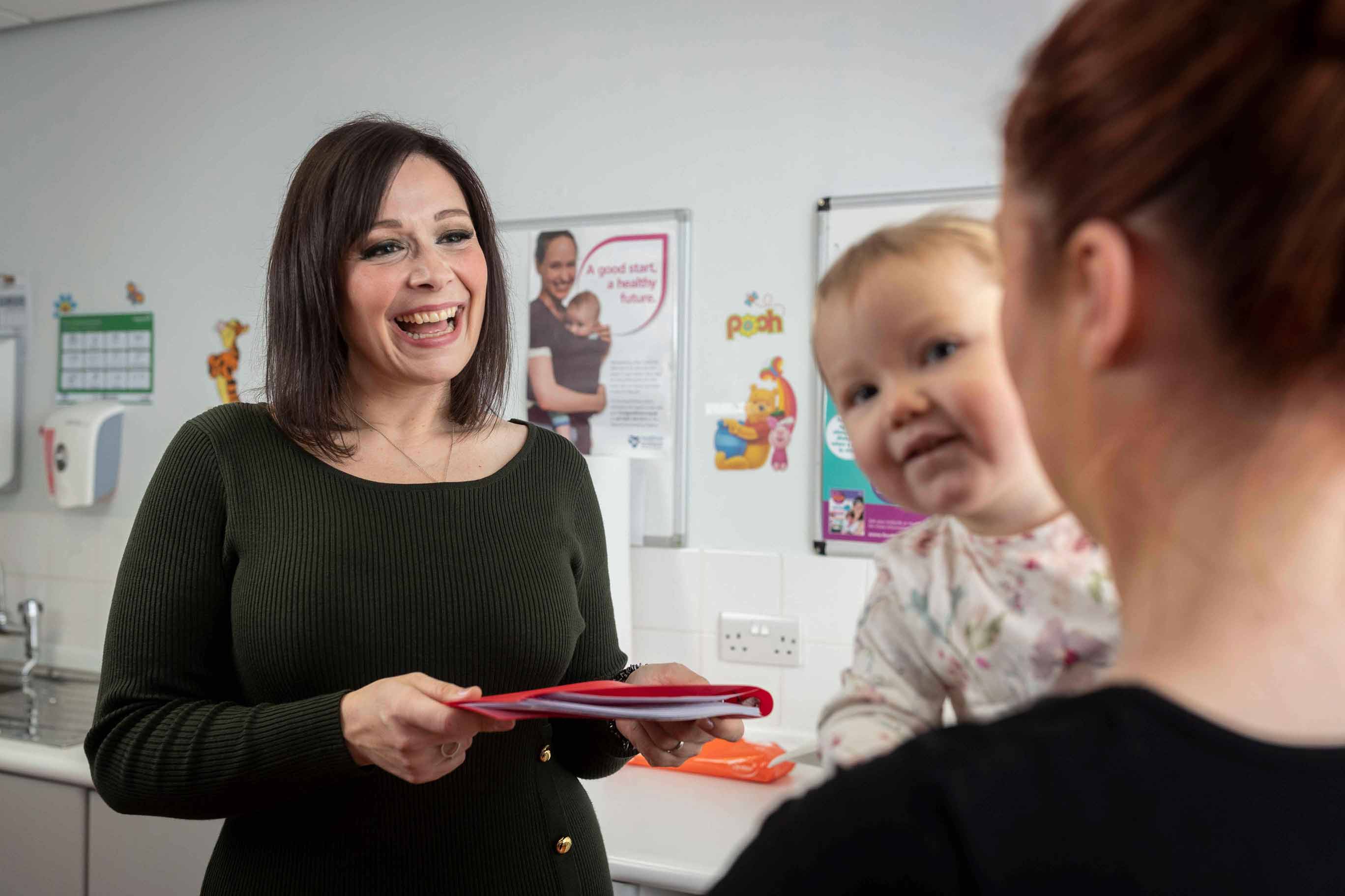 A smiling person with short dark hair, wearing a black round neck long-sleeved top and holding a red folder. They are talking to another person who has their back to the camera. They are holding a baby. In the background is a clinical office setting with a sink, soap holder and posters on the wall.