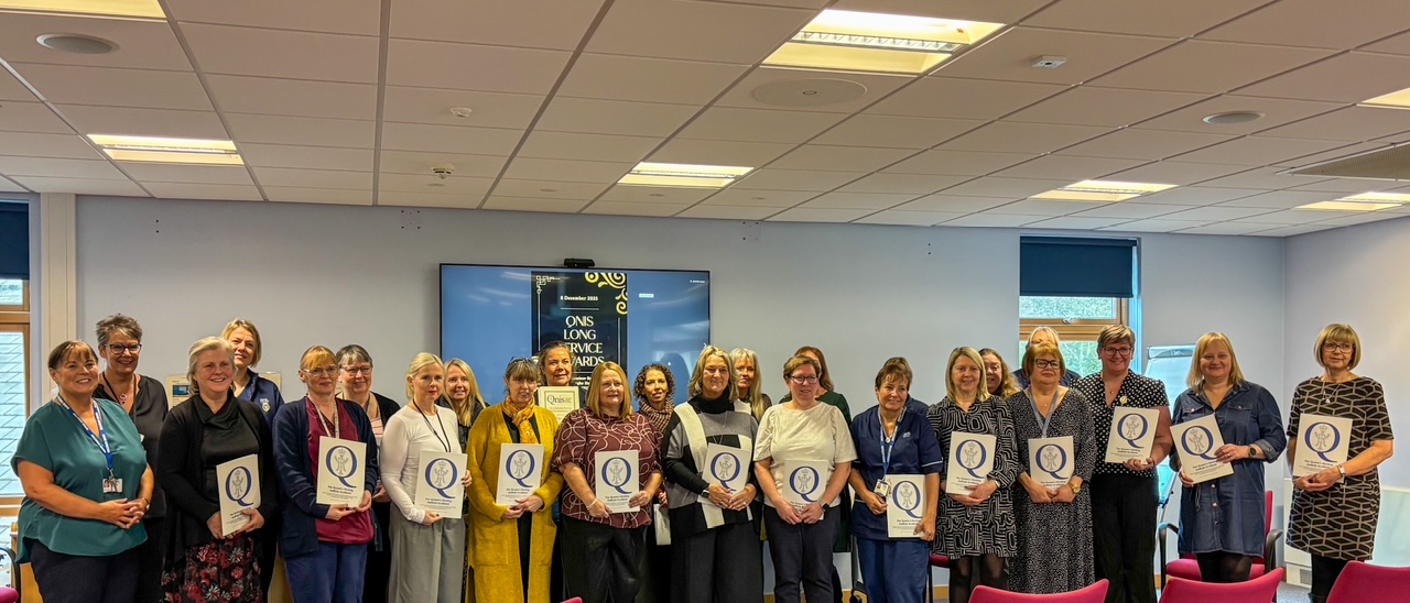 A large group of people standing in a line indoors, posing for a photo. Many are holding certificates featuring a large letter ‘Q’. Behind them is a screen displaying information about a long service awards event. The setting appears to be a professional or celebratory gathering.