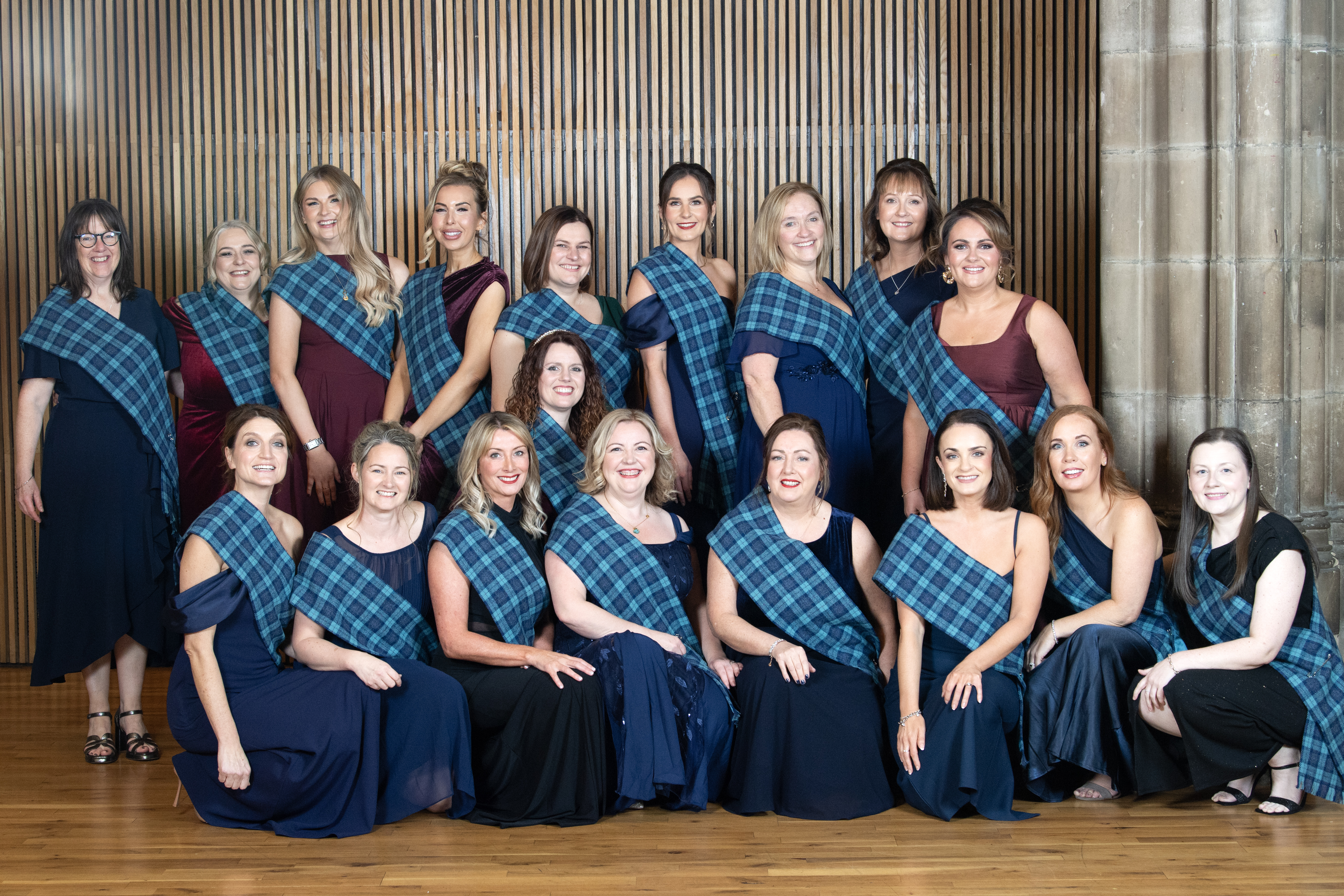 A group of people dressed in formal dark outfits with blue tartan sashes pose together indoors. They are arranged in two rows, standing and seated, in front of a wooden panelled wall and stone column.