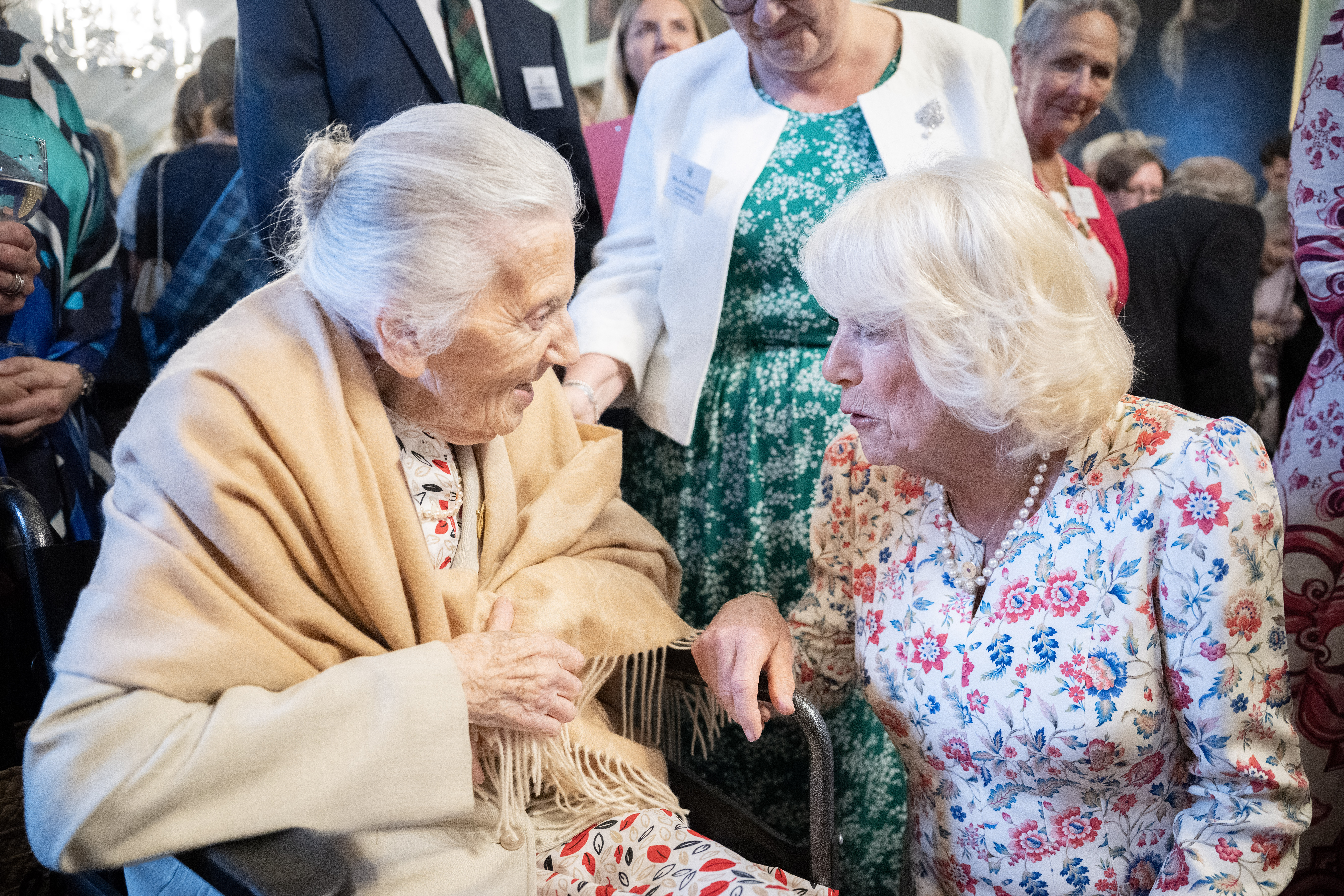 Two people are having a conversation. One is seated in a wheelchair, the other is knelt beside them. Other people look on in the background.