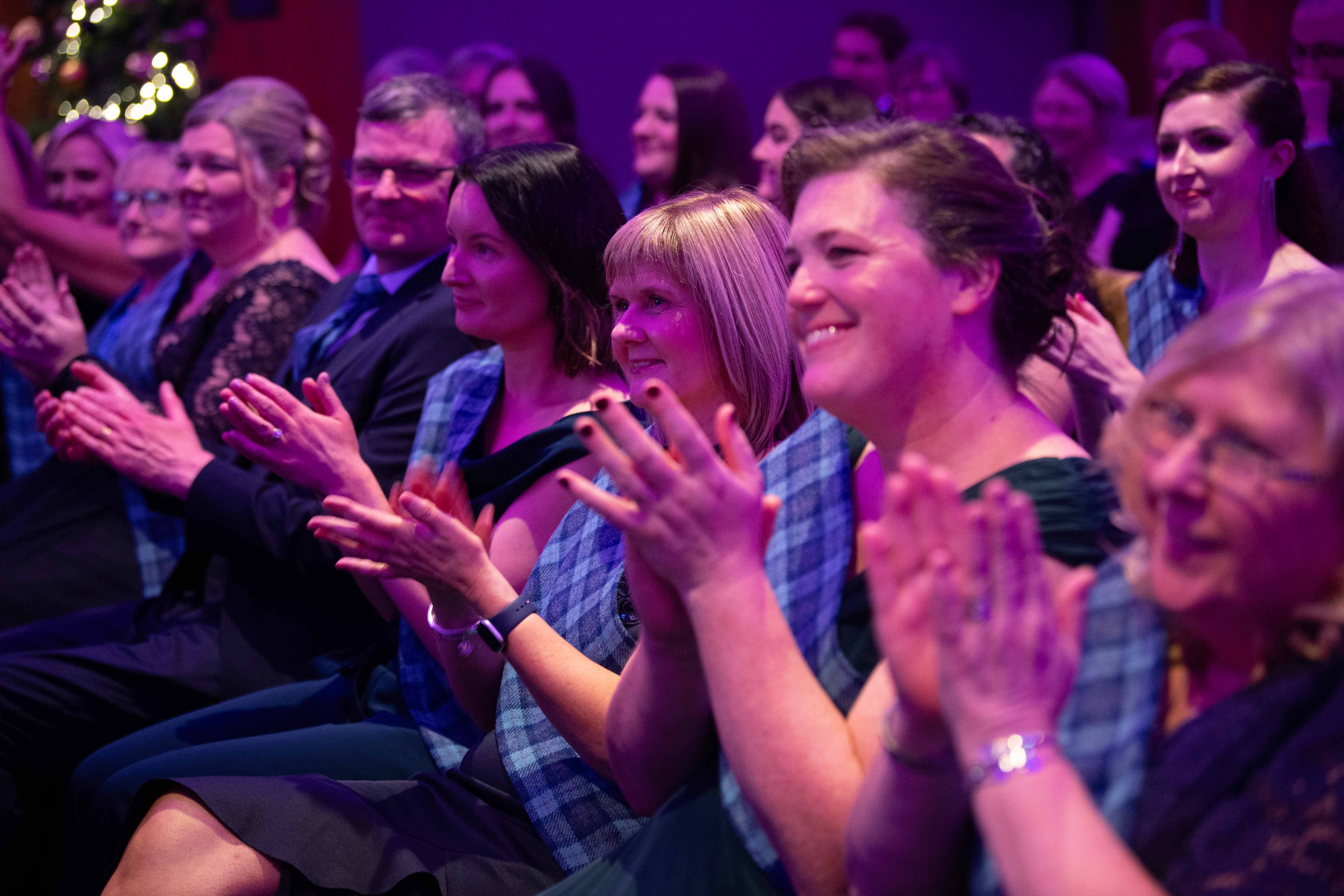 A group of people seated in rows, clapping and smiling. Some individuals are wearing tartan sashes over dark clothing. The lighting is purple and festive, and the atmosphere appears celebratory.