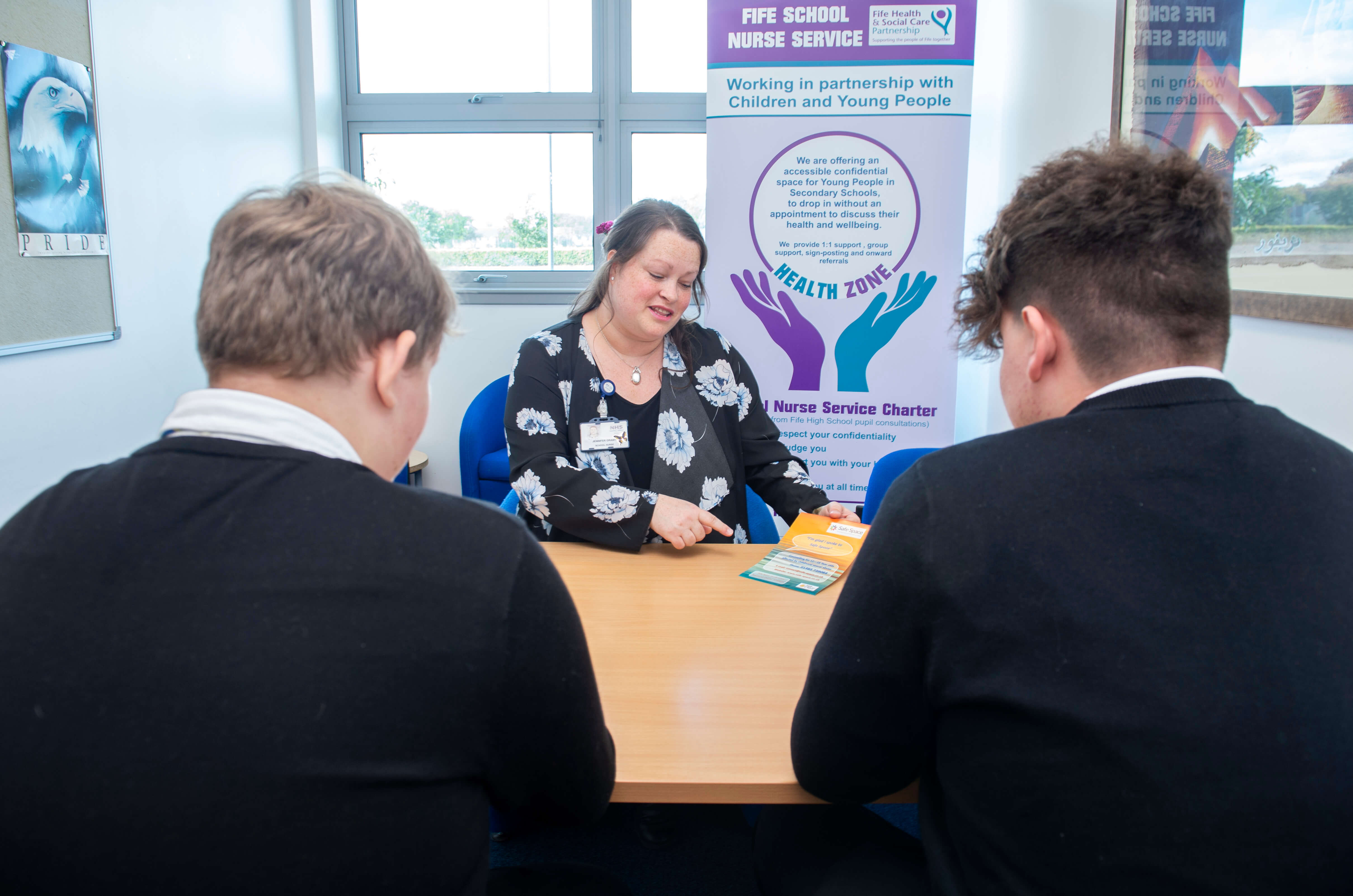 A person sits at a desk speaking to two young people across from them. The person at the desk is holding a leaflet and smiling, with a banner behind them that reads 'Fife School Nurse Service' and describes support available for children and young people. The setting appears to be a school office or consultation room.