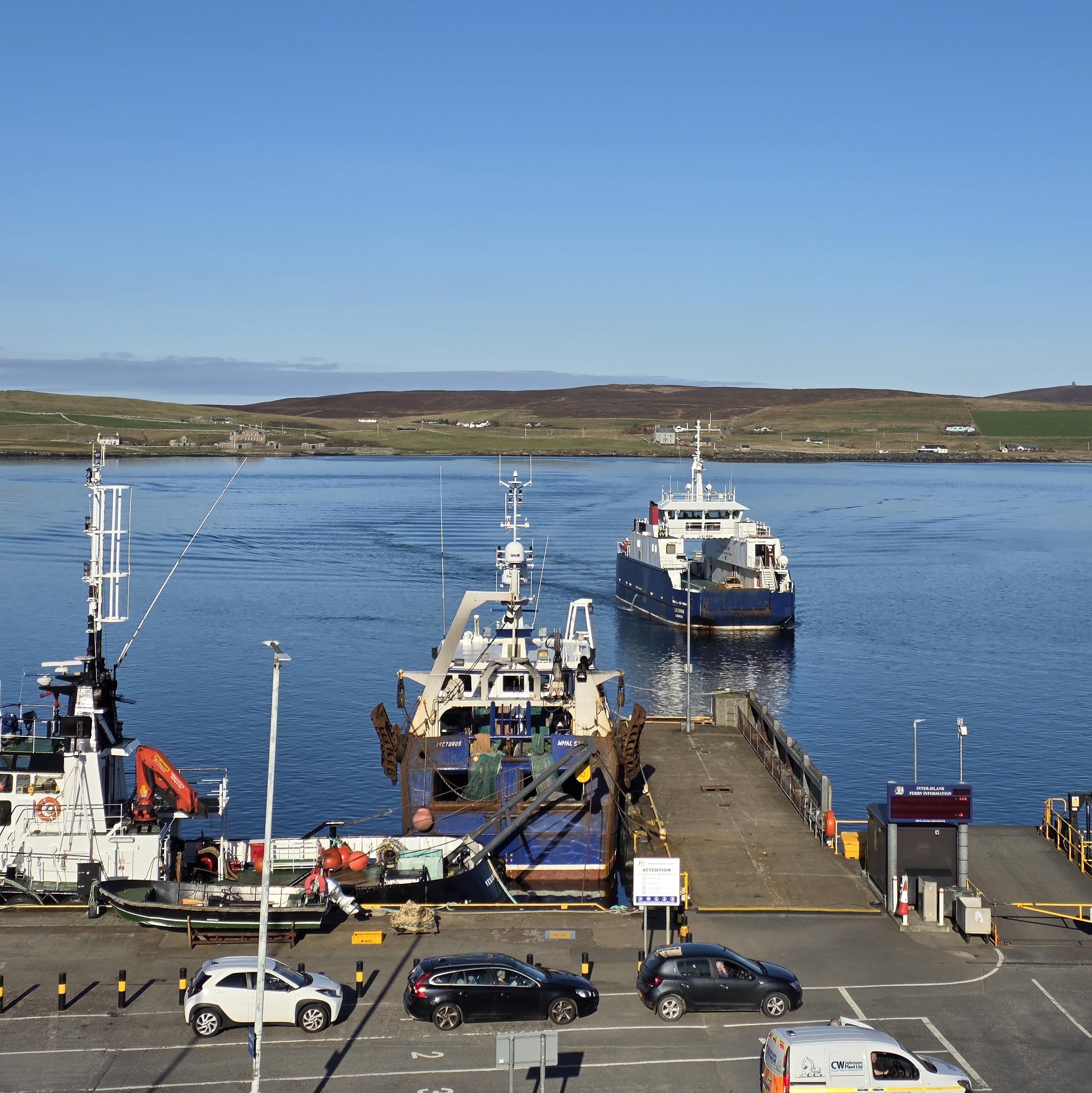 A small harbour with calm blue water under a clear sky. In the foreground, a dock holds fishing vessels and equipment, including cranes and nets, with a few parked cars along the quay. A ferry approaches the pier from the water, facing toward the dock. Across the water, low rolling hills dotted with scattered houses stretch along the shoreline.