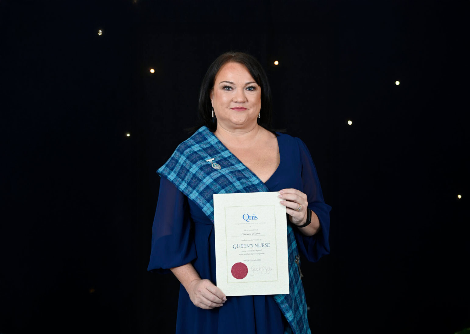 A person with straight dark hair stands in front of a dark background with small lights, wearing a blue dress and a blue tartan sash. They are holding a certificate that reads "Queen's Nurse" and smiling at the camera.