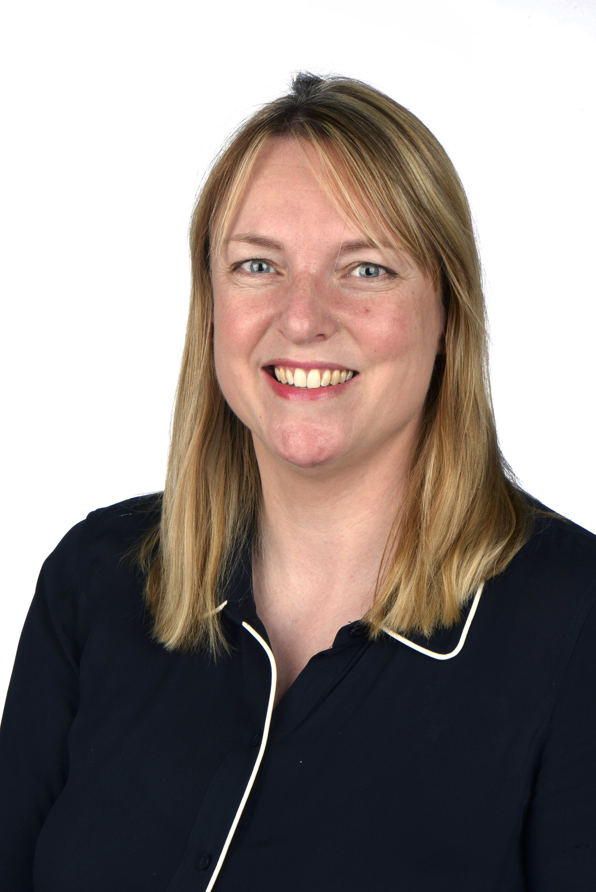 A person with straight, shoulder-length blonde hair and blue eyes is smiling at the camera. They are wearing a dark navy blouse with white piping along the collar and neckline. The background is plain white, and the photo appears to be a professional headshot.