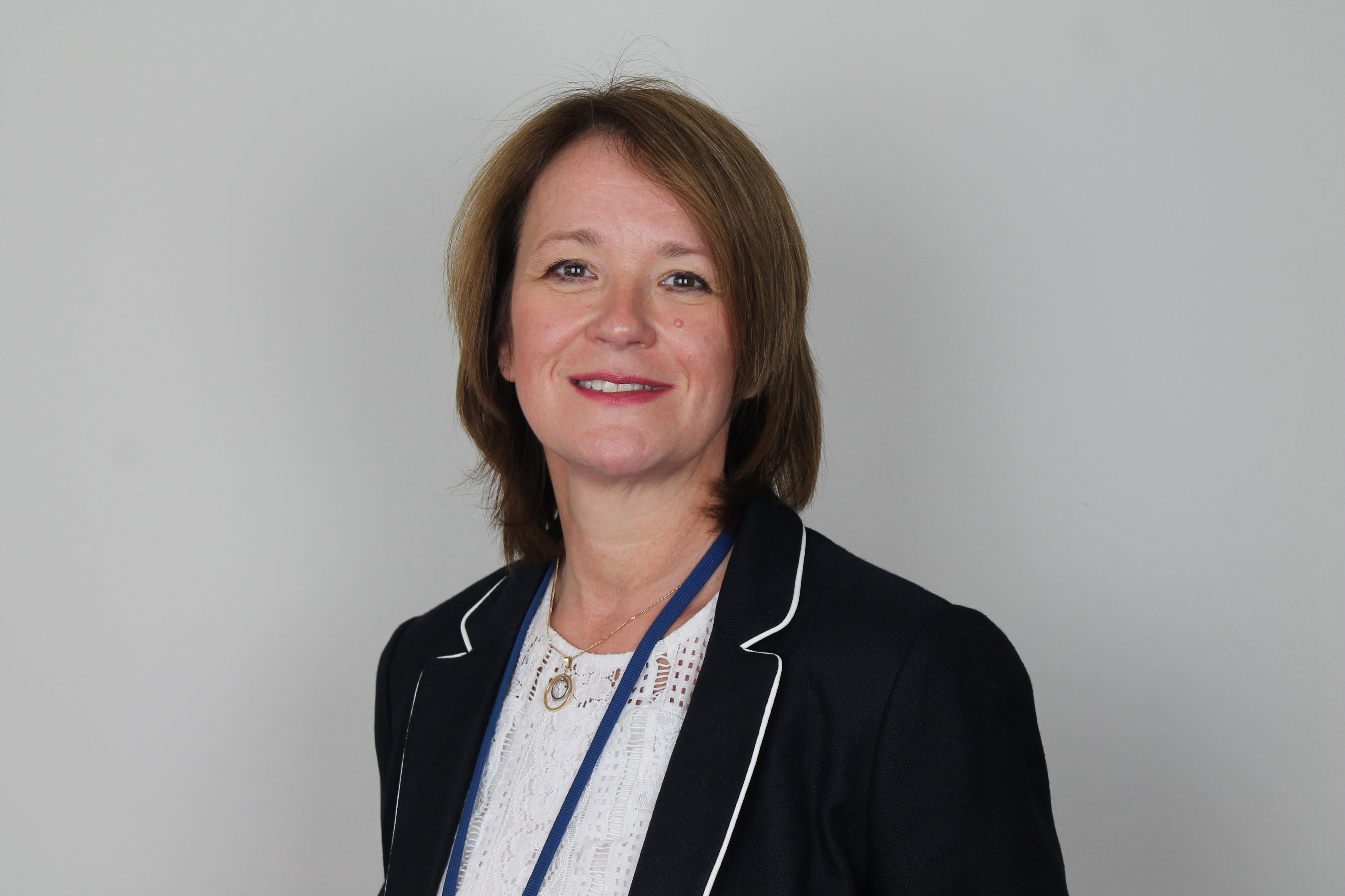 A person with short, light brown hair wearing a dark blazer with white piping, a white lace top, and a blue lanyard. They are standing in front of a plain, light gray background and smiling at the camera.