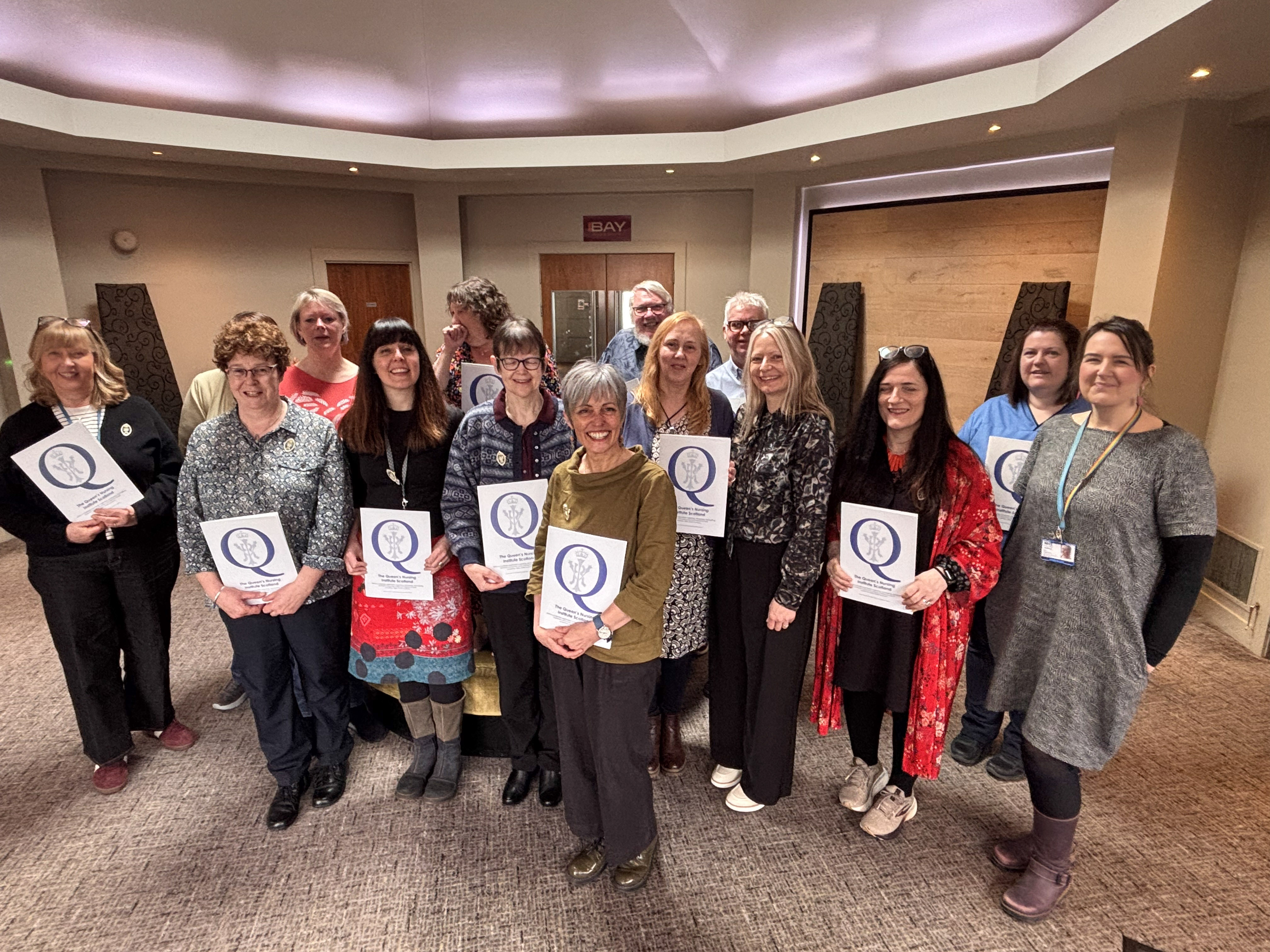 A group of 15 adults stand together in a warmly lit indoor room, smiling at the camera. Many are holding certificates with a large “Q” logo on the front. The group is arranged in two rows, dressed in casual to smart-casual clothing. The space has carpeted flooring, soft overhead lighting, and wooden wall panels, suggesting a meeting or event setting.