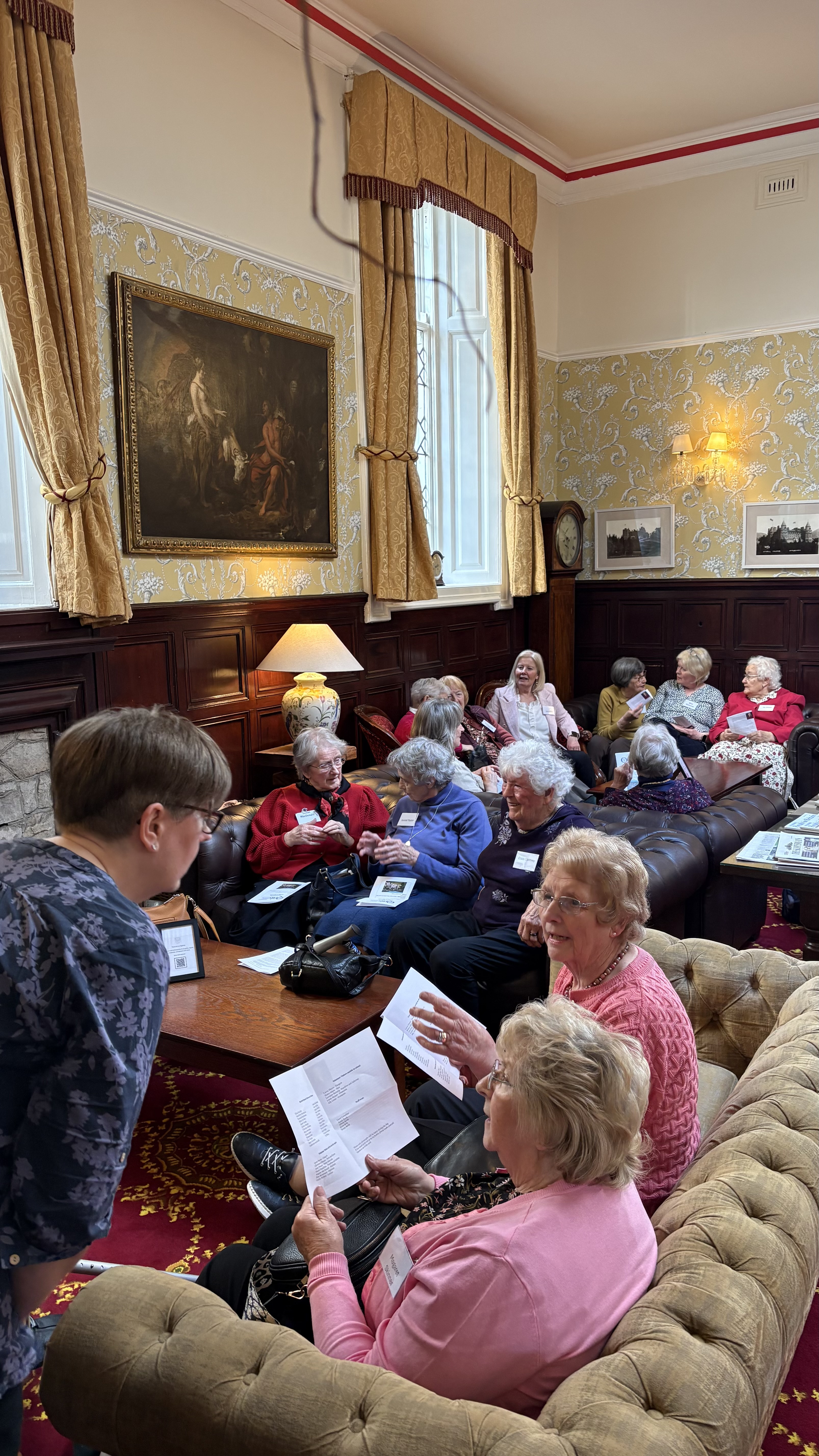 A group of older adults sit and chat in a comfortable, traditionally decorated lounge with patterned wallpaper, tall windows, and framed artwork. Several people hold printed papers and appear to be discussing or reading together. One person leans in from the foreground to speak with others seated on sofas and chairs, creating a relaxed, social atmosphere.