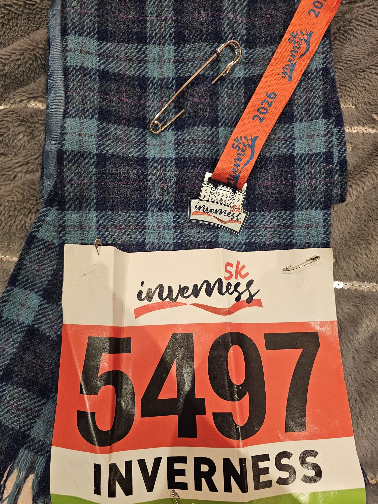 The image shows a runner's bib displaying the number 5497 which is placed on top of a tartan sash