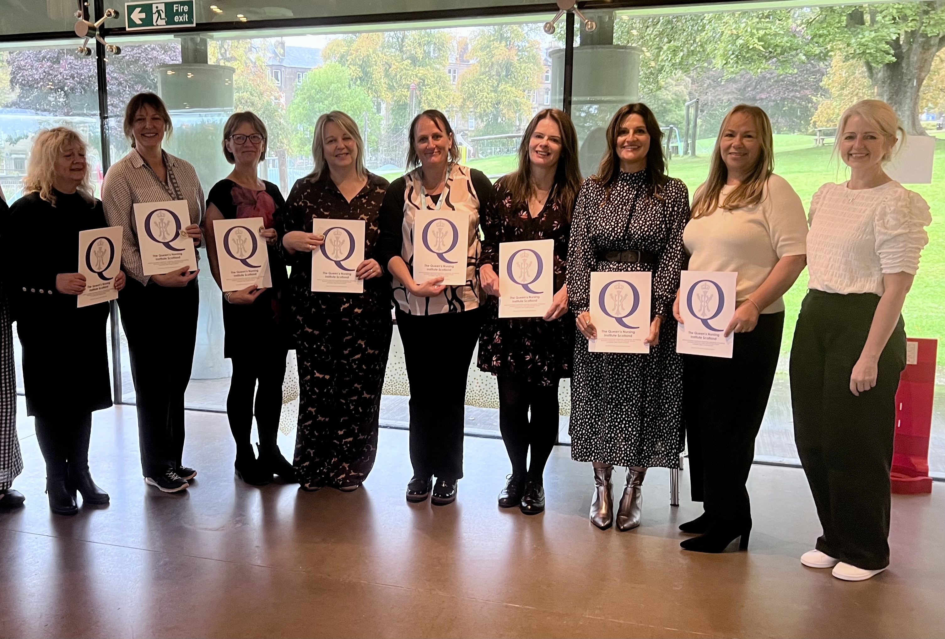 A group of people standing in a line indoors in front of large windows overlooking a green outdoor area. Several individuals are holding certificates featuring a large letter ‘Q’. Everyone is posing for a group photo at what appears to be a professional or recognition event.