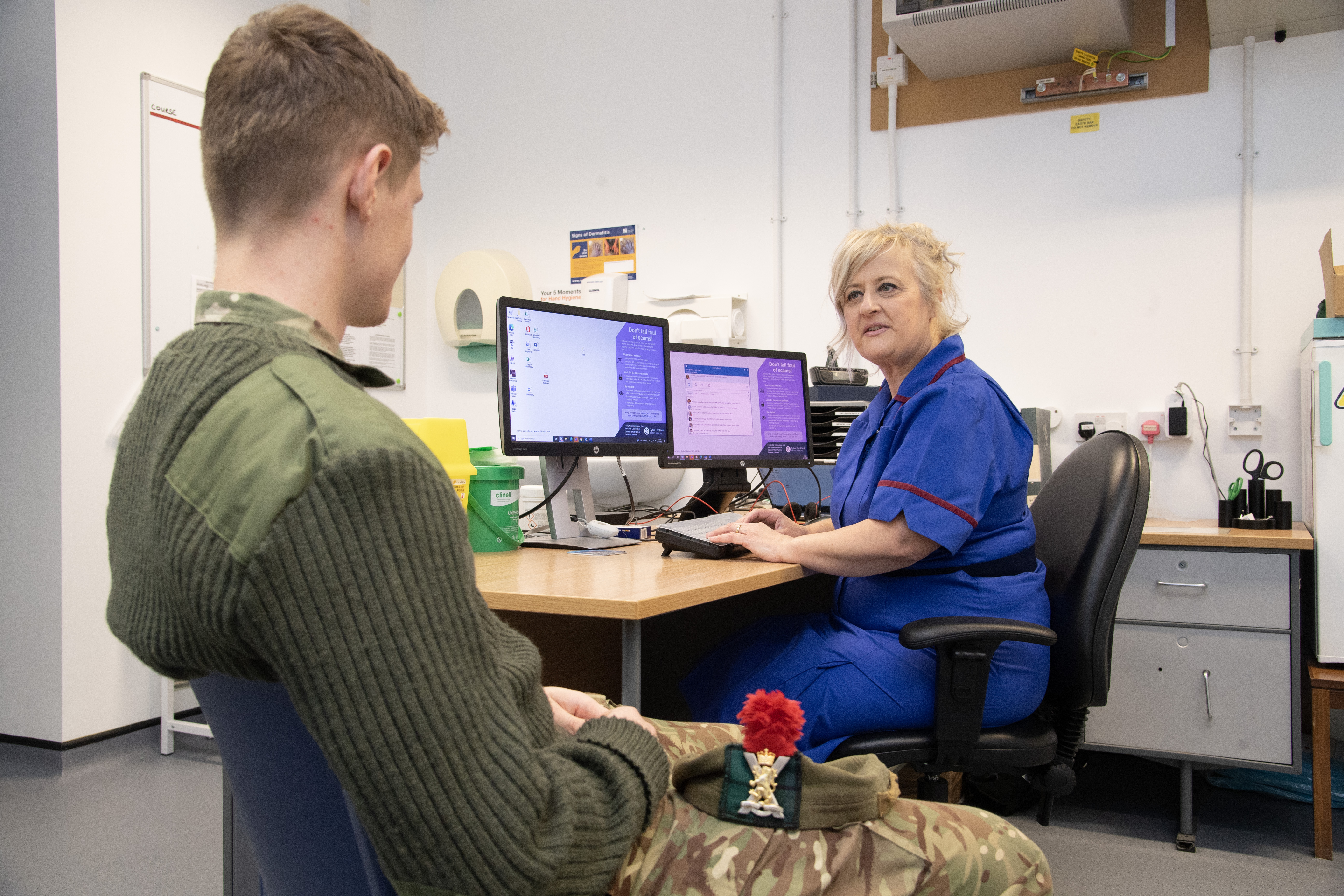 Two people sit across from each other next to a desk with two computer monitors on it. The person with their back to the camera has short light brown hair and is wearing a green army uniform, jumper, camouflage trousers and a beret with a badge, tartan and a red plume. The person facing the camera has tied back blonde hair and wears a blue nurse’s uniform. In the background are drawers, a grey floor and white walls. A fuse box is just visible on the wall.