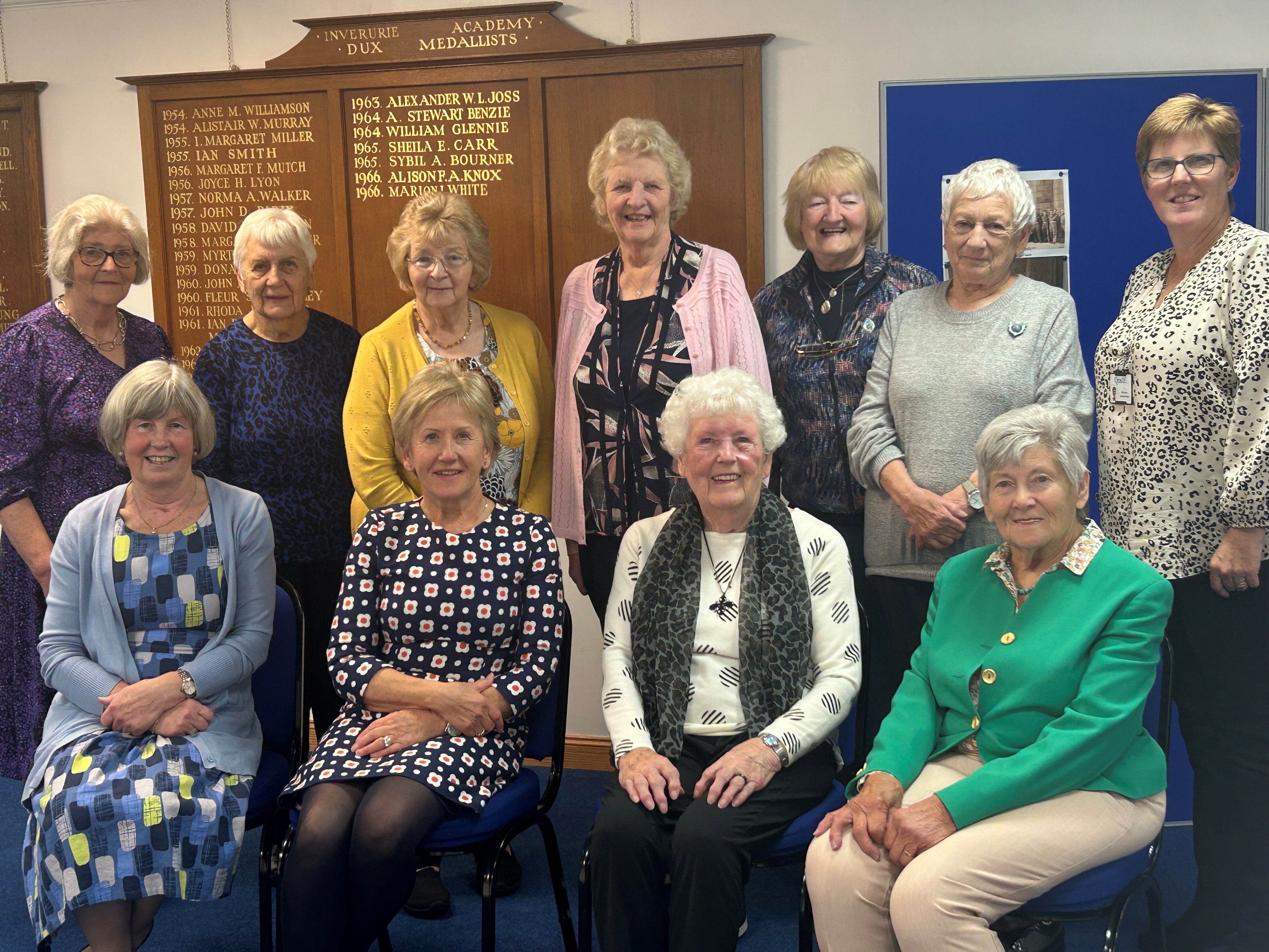 A colour image of a group of older adults smiling and seated and standing together. A wooden wall plaque with gold lettering saying Inverurie Academy Dux Medallists is behind them.