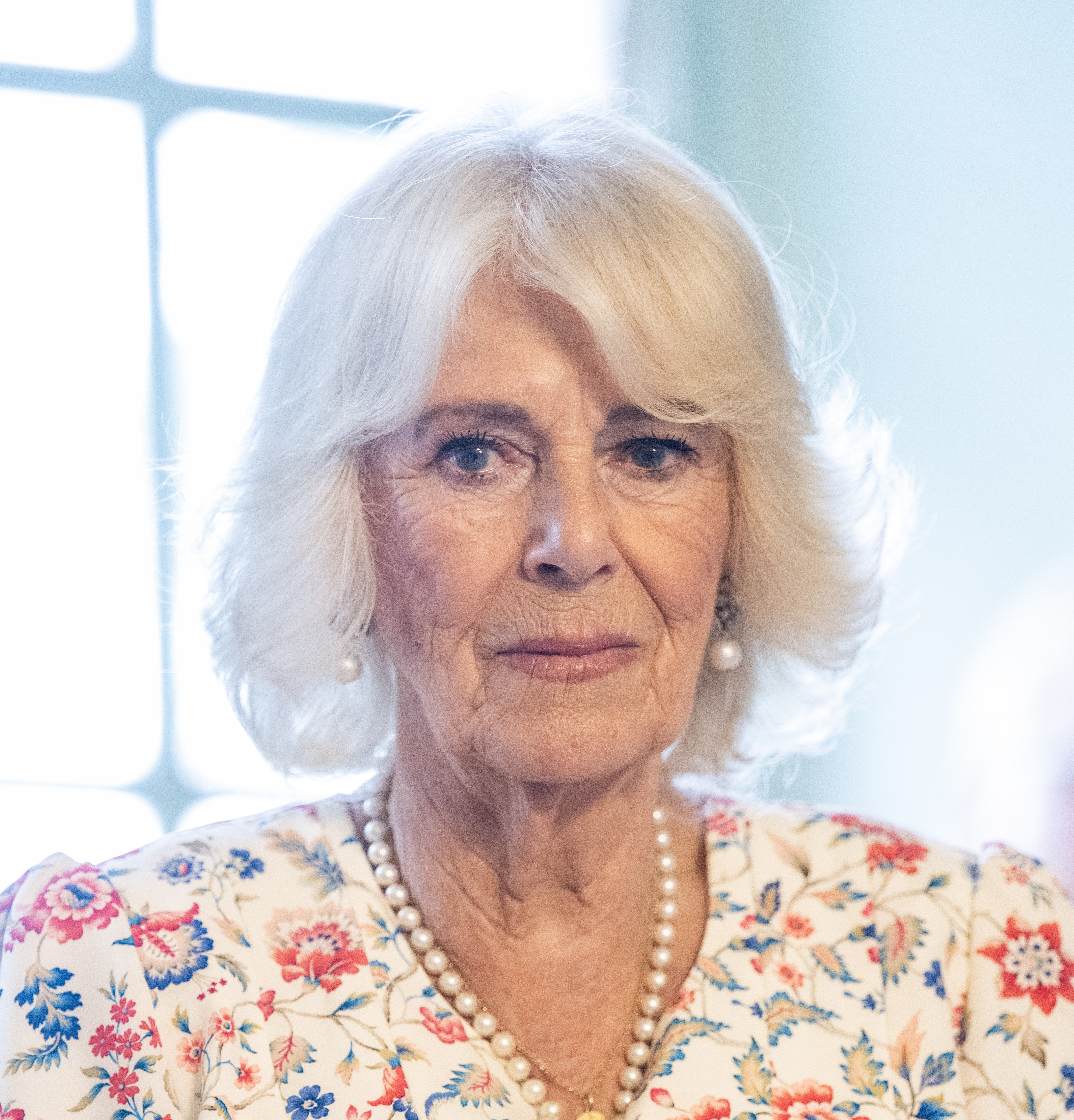 A person with shoulder-length pale blonde hair stands indoors in soft natural light. They are wearing a floral-patterned outfit with red, blue, and cream tones, a pearl necklace, and matching pearl earrings. Their expression is neutral, and the background features a softly lit window.