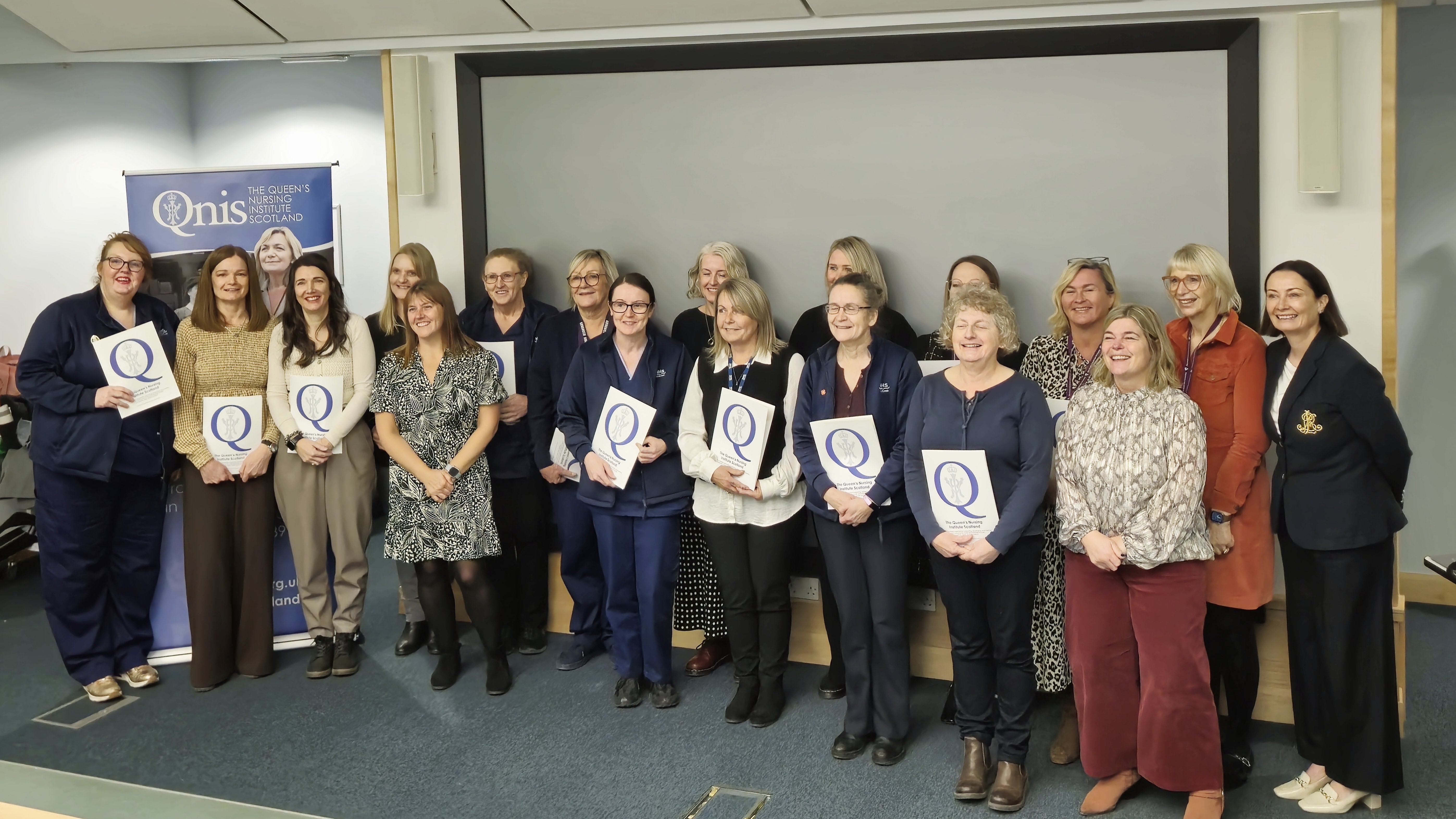 A group of people standing together indoors in front of a presentation screen and a banner for the Queen’s Nursing Institute Scotland. Several individuals hold certificates featuring a large letter ‘Q’. The group is arranged in two rows, smiling and posing for a photo. The setting appears to be a formal or professional event.