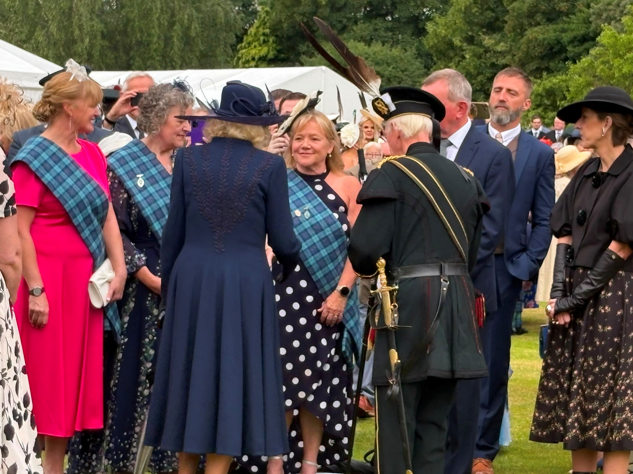 A group of people in formal attire stand together in a garden Three of the people are wearing tartan sashes.. They are chatting and smiling.