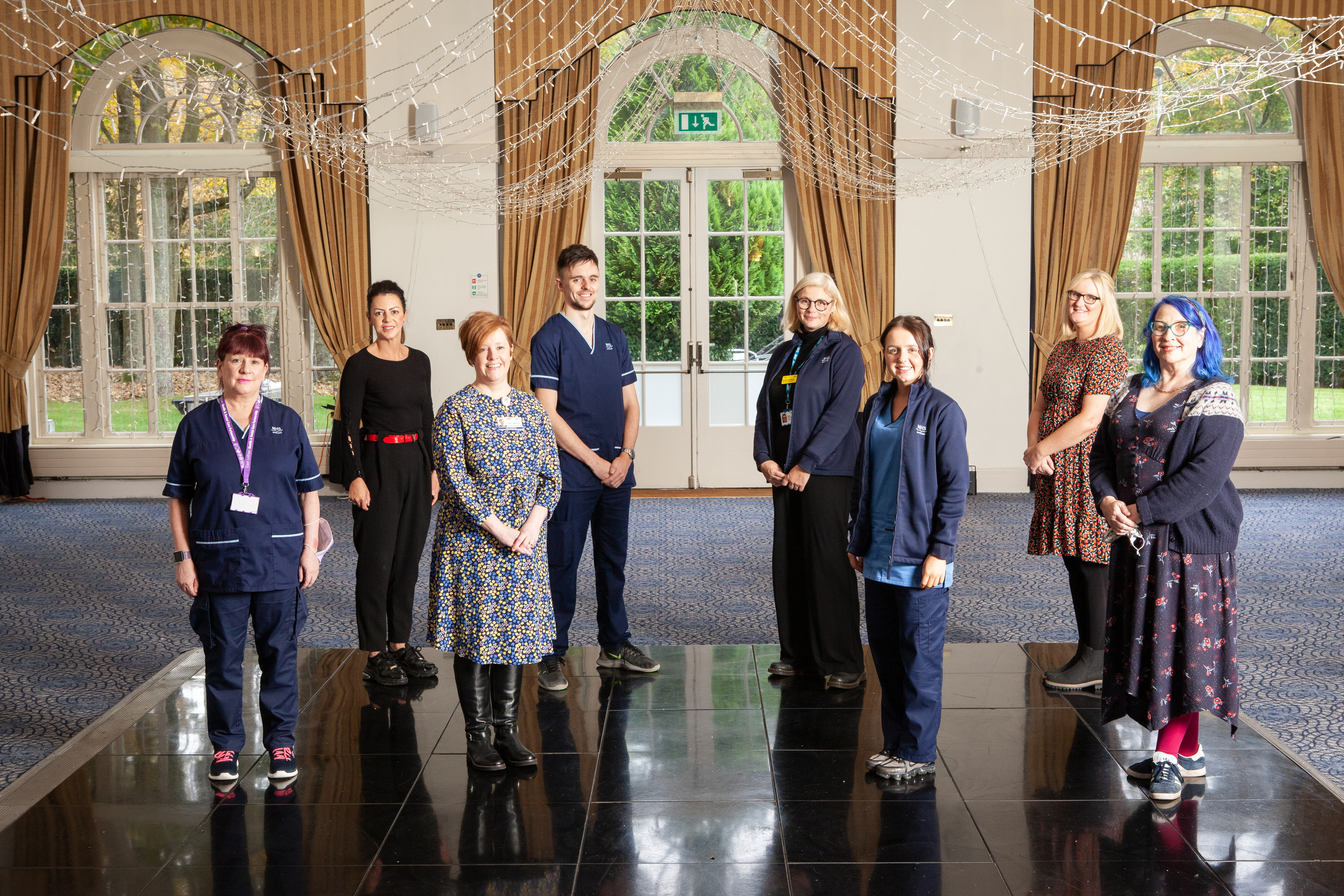 A group of 8 people stand spaced apart in twos and threes indoors in a formal room with tall arched windows, gold curtains, a blue patterned carpet and black shiny tiles, which they are standing on. Decorative string lights hang from the ceiling. The group includes individuals in professional navy uniforms, and others in casual clothing. They smiling at the camera. Trees and greenery are visible through the large windows in the background.